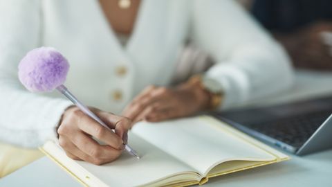 Close-up of a woman writing notes with a fluffy pen, indoors near a laptop.