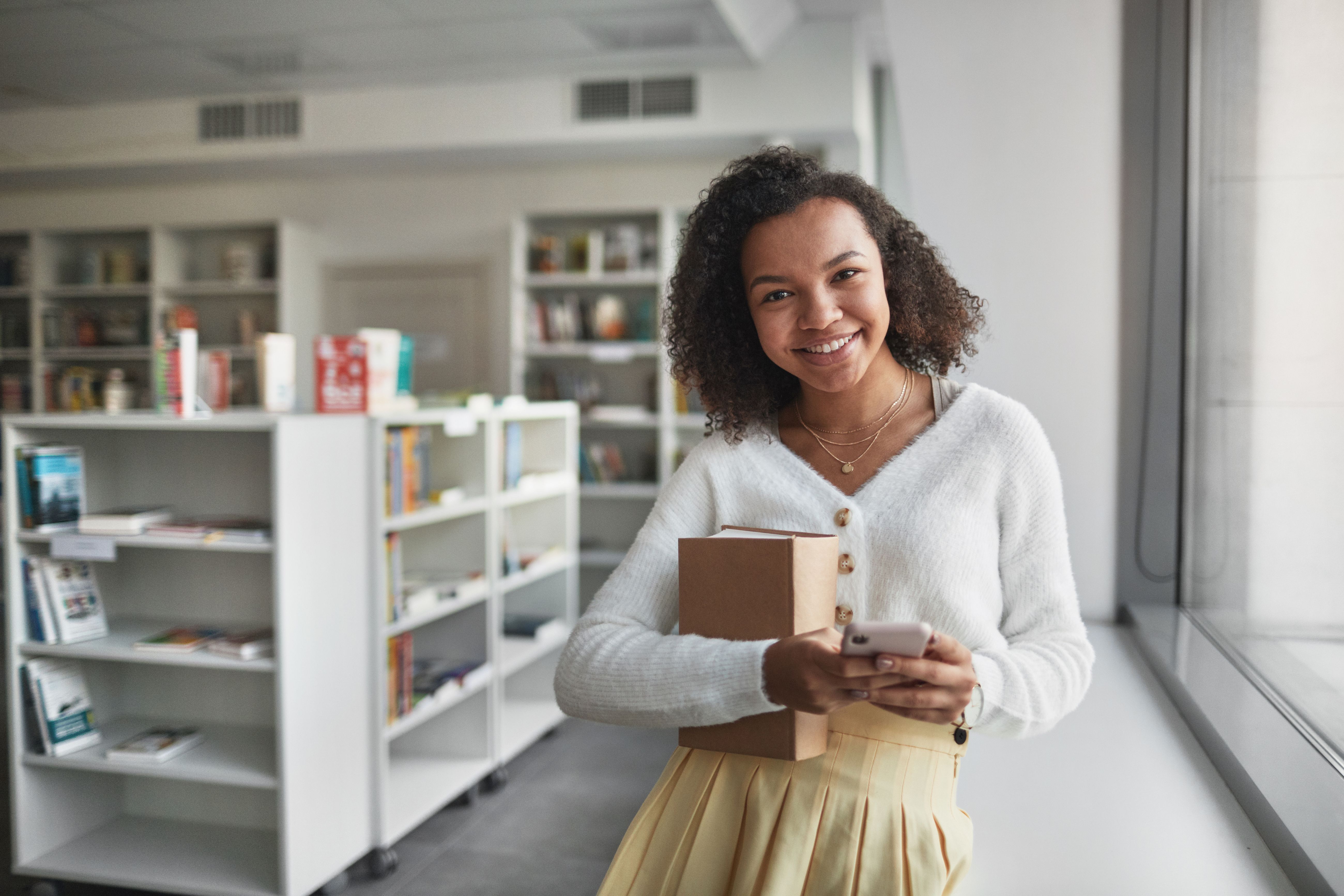 Confident black woman in a modern library holding a book and smartphone, smiling warmly.