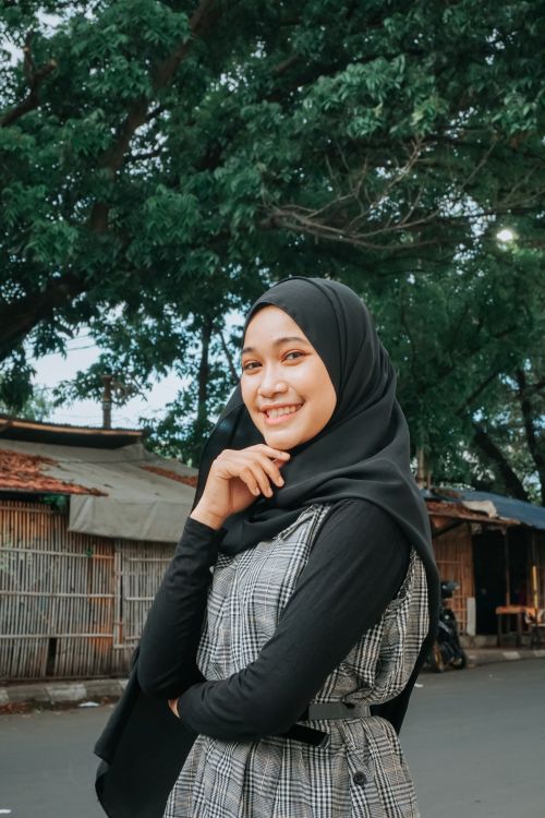 Free Portrait of a smiling woman in a hijab posing in an outdoor setting with trees and buildings in the background. Stock Photo