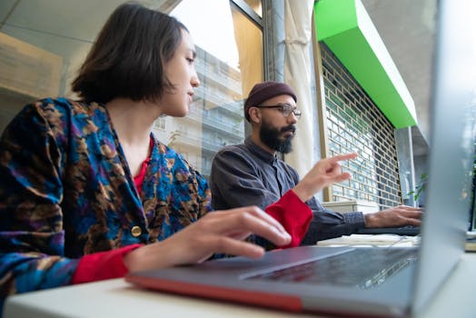 Two professionals collaborating at a laptop in a contemporary glass-walled office setting.