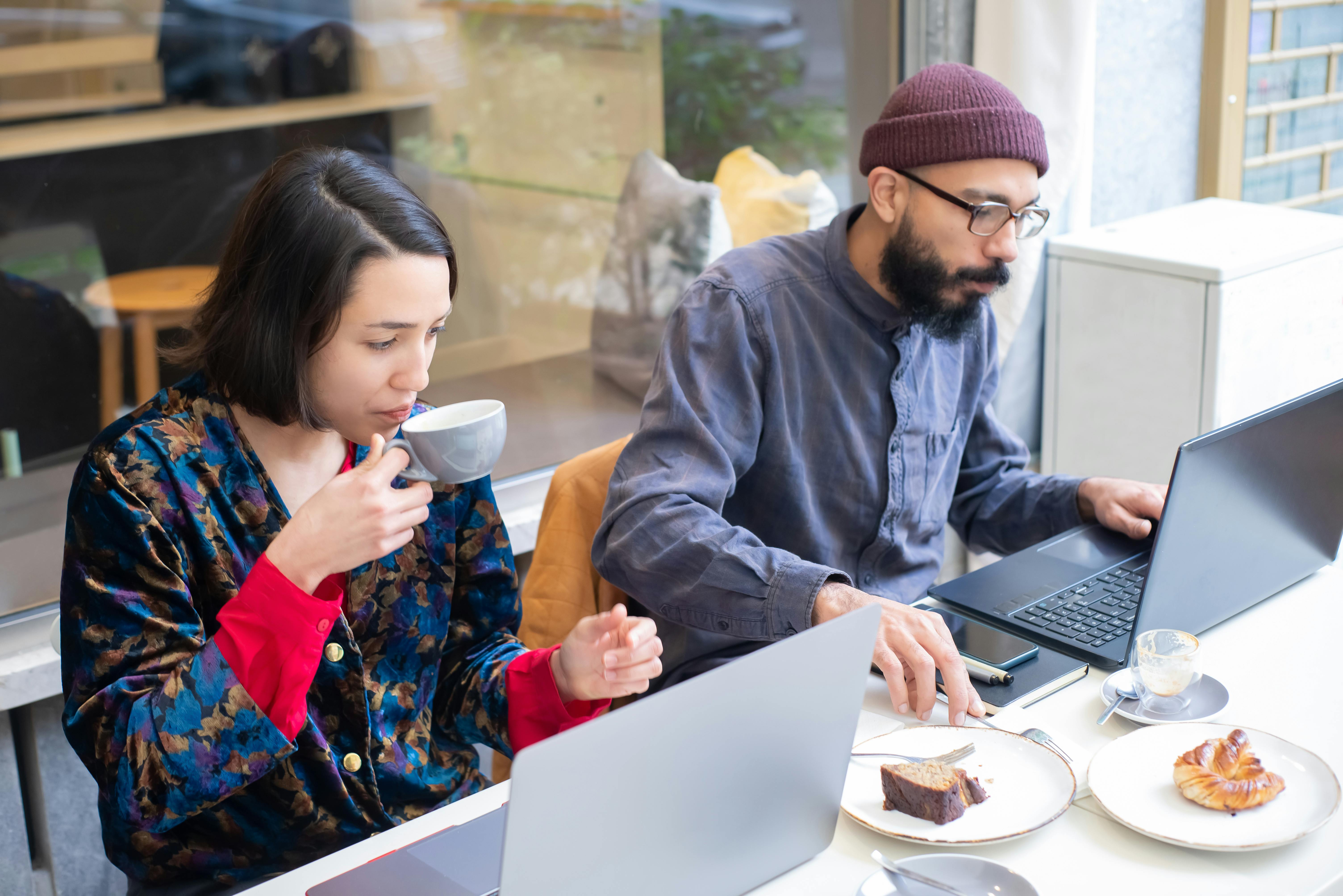 Two adults working on laptops in a contemporary café, enjoying coffee and pastries.