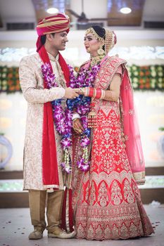 Full body of smiling Indian couple in traditional clothes standing in decorated hall and holding hands while looking at each other on wedding ceremony