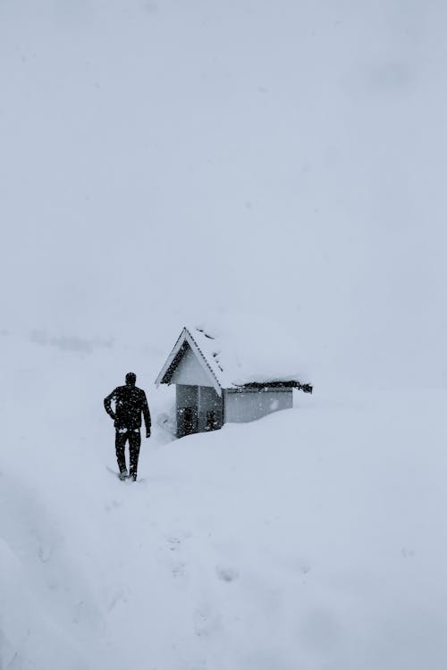 Full body back view of unrecognizable male in outerwear walking towards hut during snowfall in overcast winter day