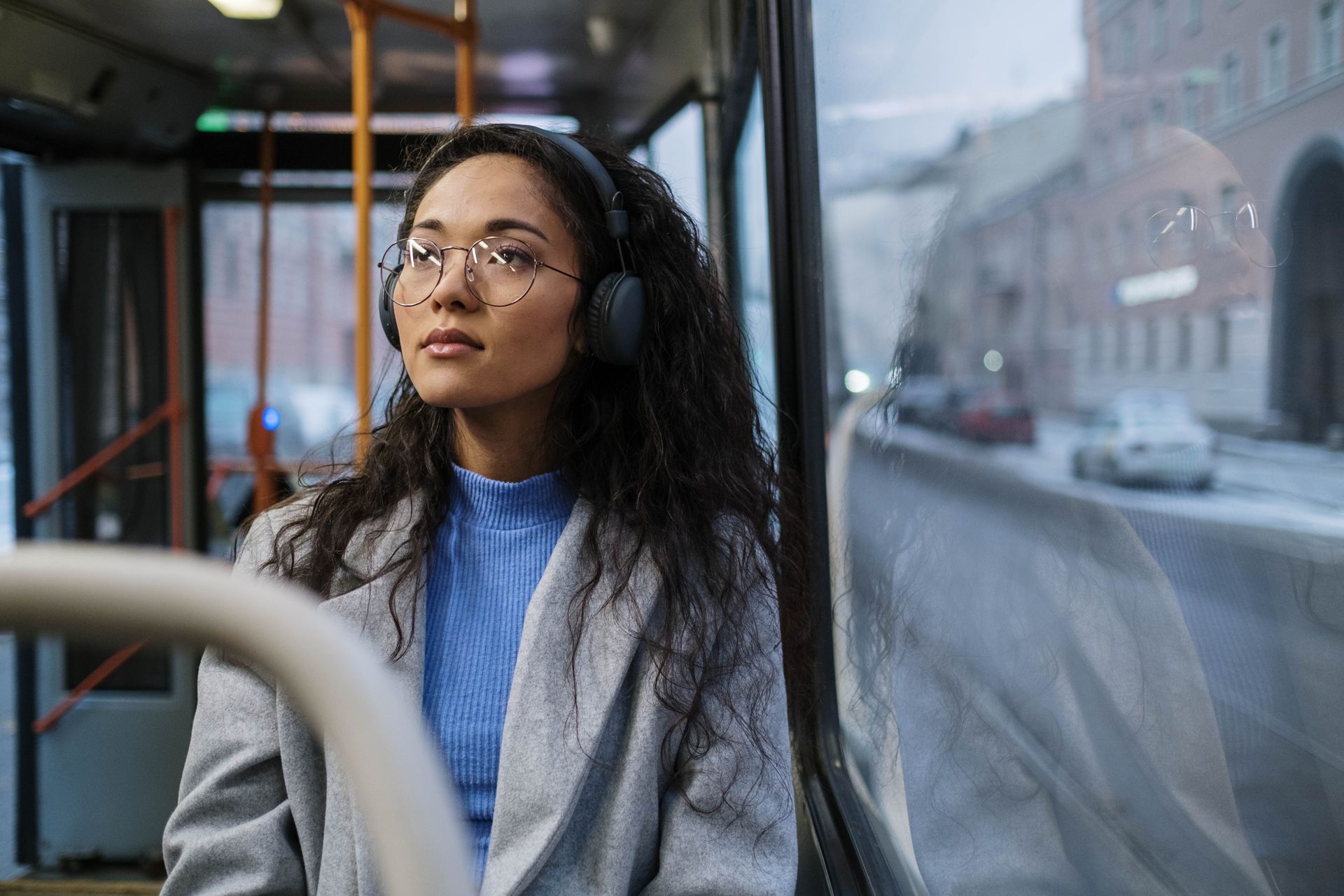 Young woman gazing out bus window, wearing headphones and glasses.