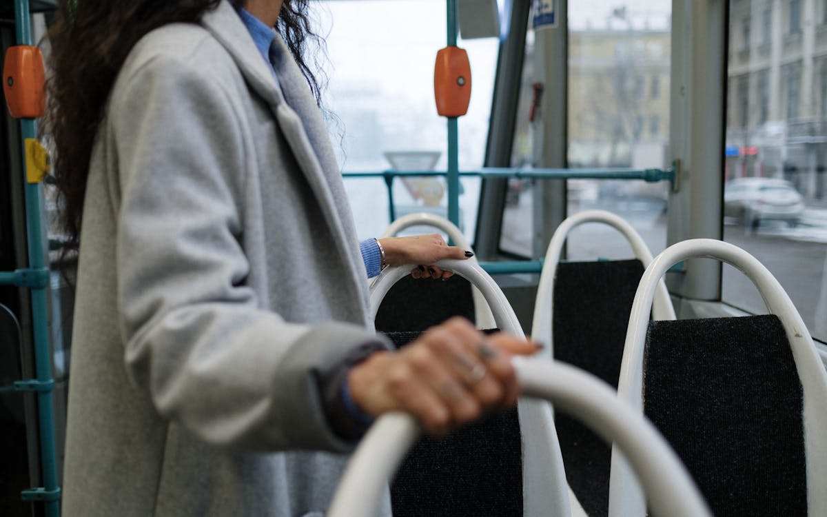 A person holding a rail inside a city bus, wearing a gray coat, with a street view outside.