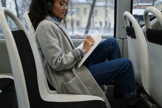 Woman writes in a notebook while listening to music on a city bus.