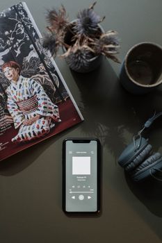 A flat lay of a smartphone showing a podcast, headphones, a magazine, and a cup on a table with soft lighting.