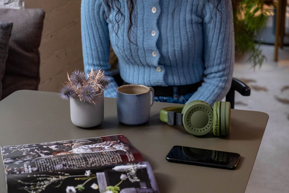 A person in a blue sweater sits at a table with headphones, a phone, and a mug indoors.