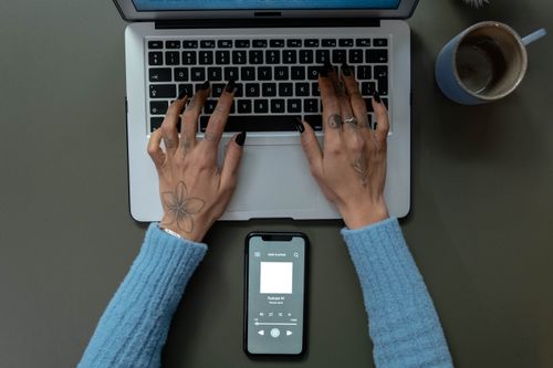 Free Top-down shot of a woman typing on a laptop with a smartphone displaying a podcast and a mug on the side. Stock Photo