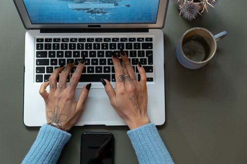 Free Overhead view of tattooed hands typing on a laptop with a mobile phone and coffee mug nearby. Stock Photo