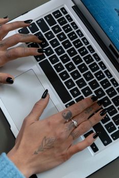 Close-up of tattooed hands with manicured nails typing on a laptop keyboard indoors.