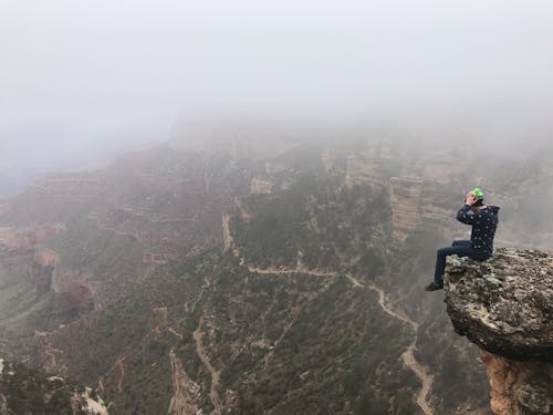 Man Sitting on Top of Cliff on Foggy Mountain