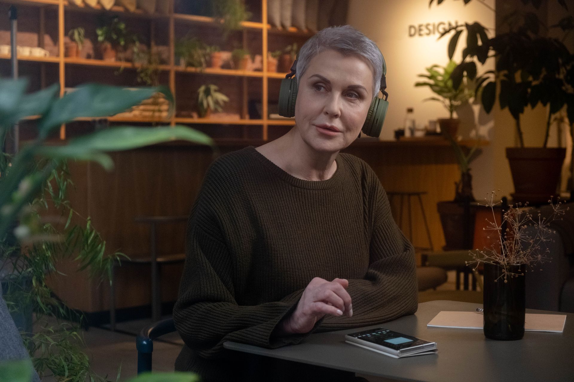 Woman enjoying music with headphones in a cozy plant-filled café interior.