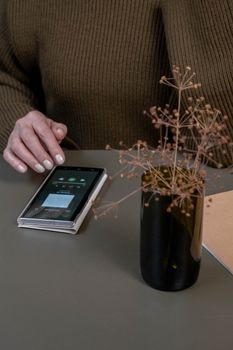 A hand touches a smartphone beside a vase with dried flowers on a table in a cozy indoor setting.