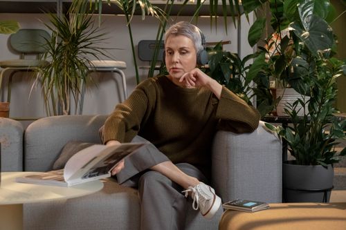 Free Mature woman sitting on sofa, enjoying music with wireless headphones, surrounded by lush houseplants. Stock Photo