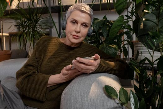 Adult woman in green sweater enjoying music with headphones, surrounded by houseplants indoors.