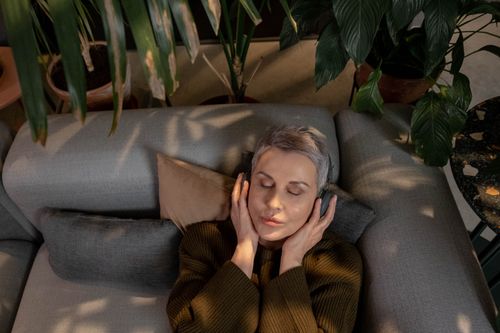 Free Mature woman with headphones relaxing on a sofa surrounded by indoor plants. Stock Photo