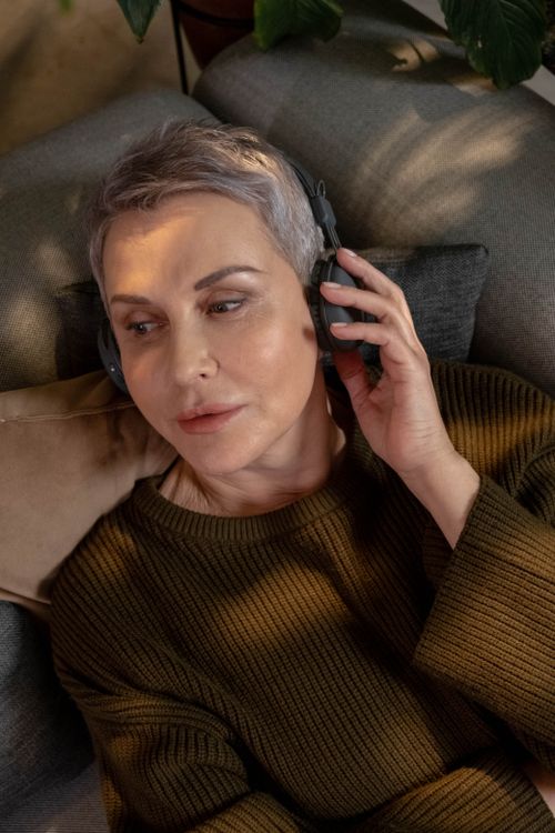 Free A senior woman with gray hair relaxes on a sofa, enjoying music through wireless headphones. Stock Photo