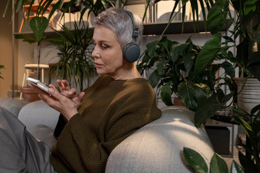 Senior woman in cozy living room using smartphone with headphones, surrounded by plants.