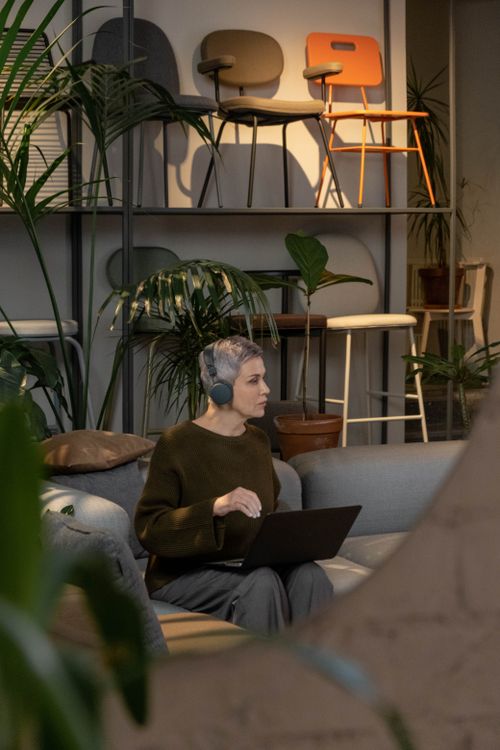 Free Mature woman sitting with laptop and headphones in a stylish indoor workspace, surrounded by plants. Stock Photo