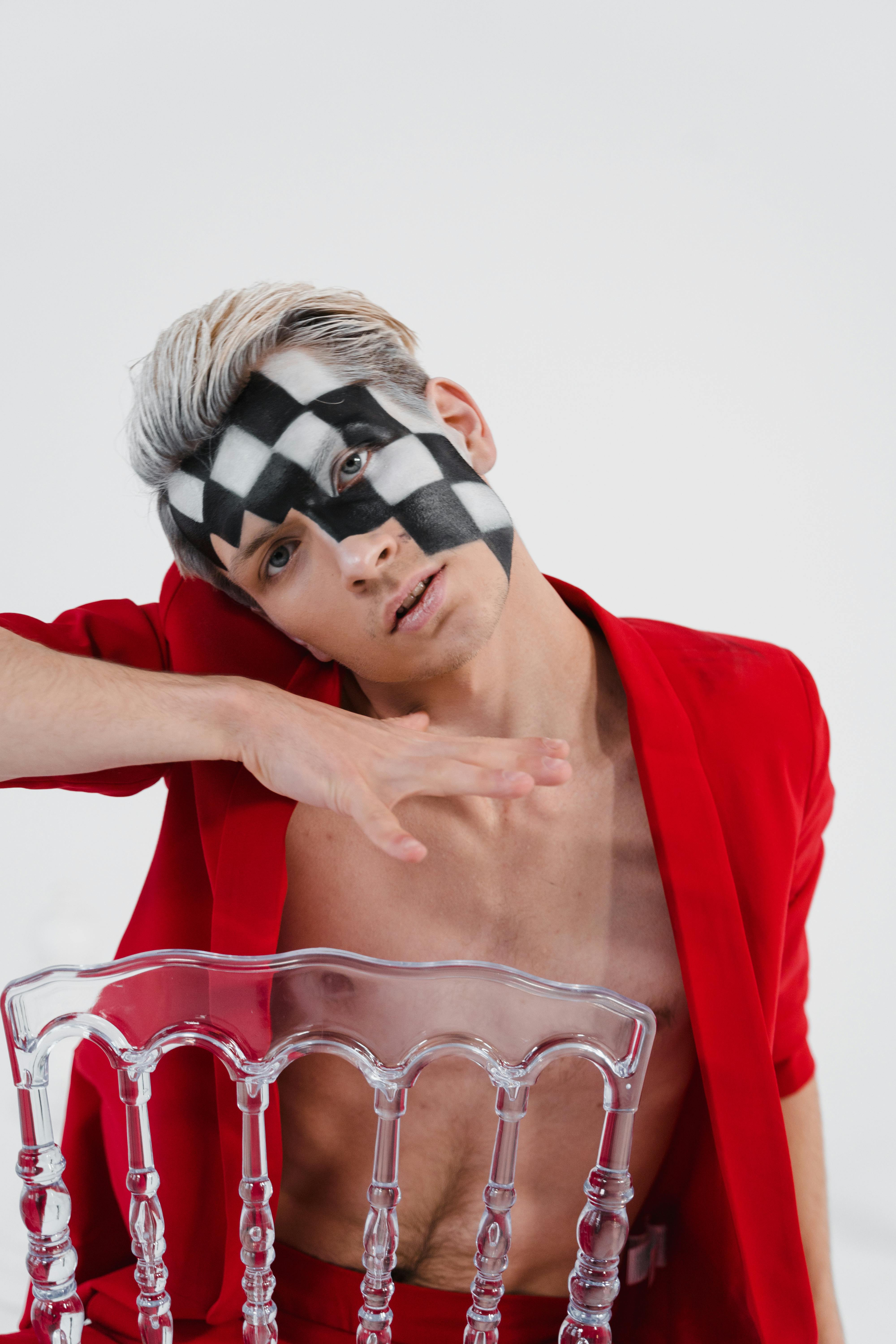 Free Artistic portrait of a man in red suit with checkerboard face paint, posing with acrylic chair. Stock Photo