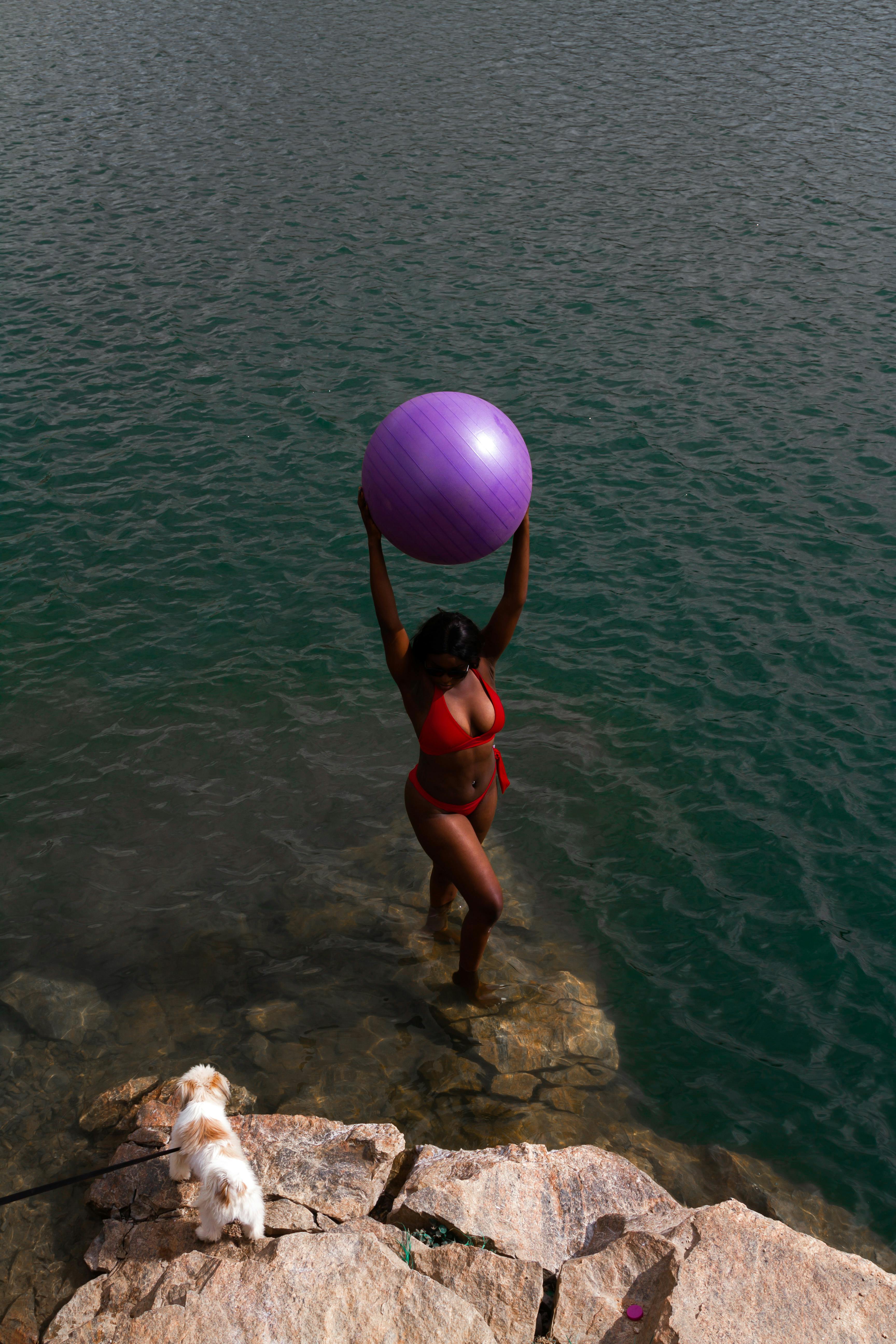 Drone view full body of unrecognizable female in bikini standing with big purple gymnastic ball on stony coast with dog near rippled ocean in daylight