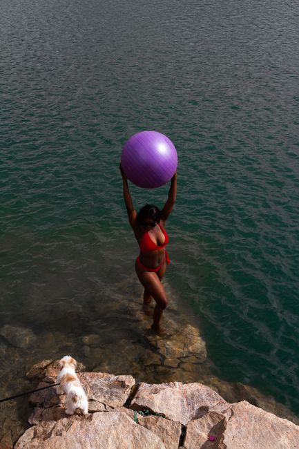 Drone view full body of unrecognizable female in bikini standing with big purple gymnastic ball on stony coast with dog near rippled ocean in daylight