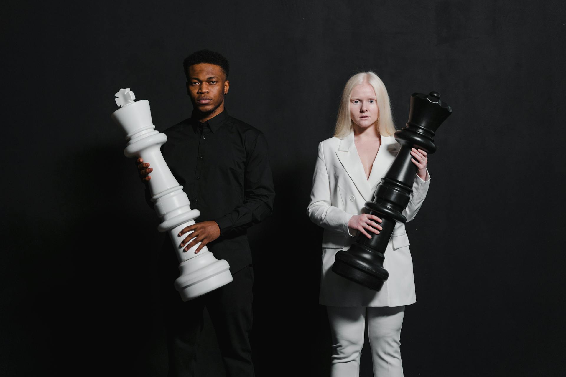 A man and woman holding oversized chess pieces against a black background.