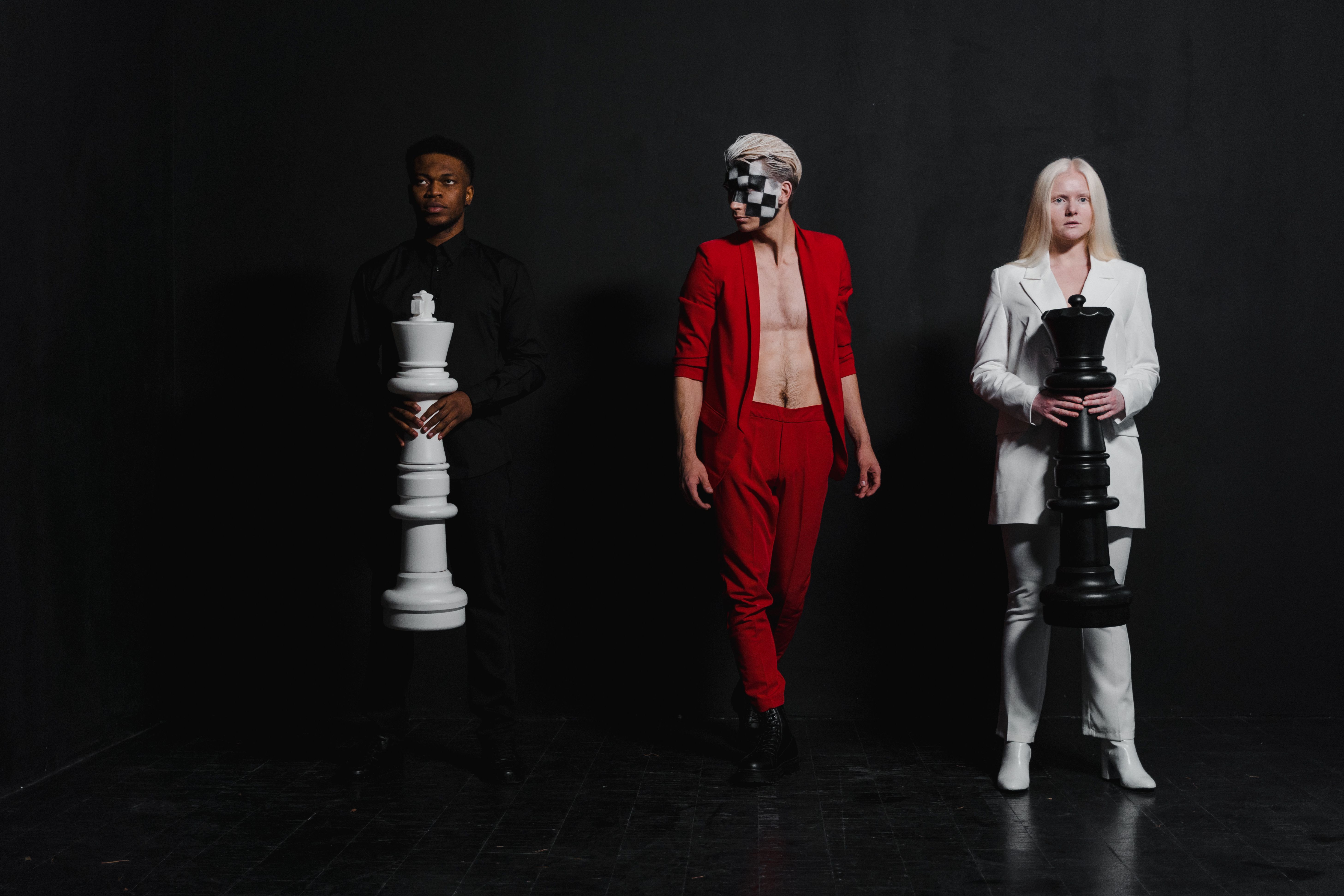 Three diverse models pose with oversized chess pieces in a conceptual studio shoot, highlighting fashion and strategy.
