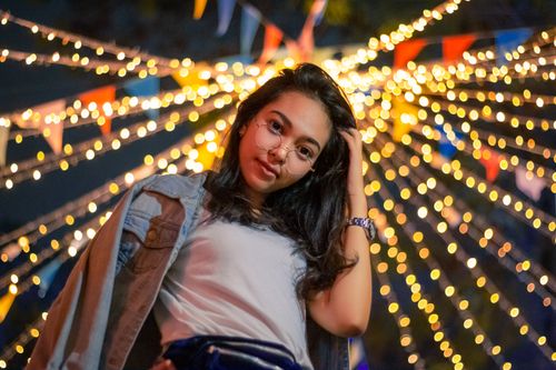 Free Stylish young woman with glasses posing outdoors at night under festive string lights in Jakarta, Indonesia. Stock Photo