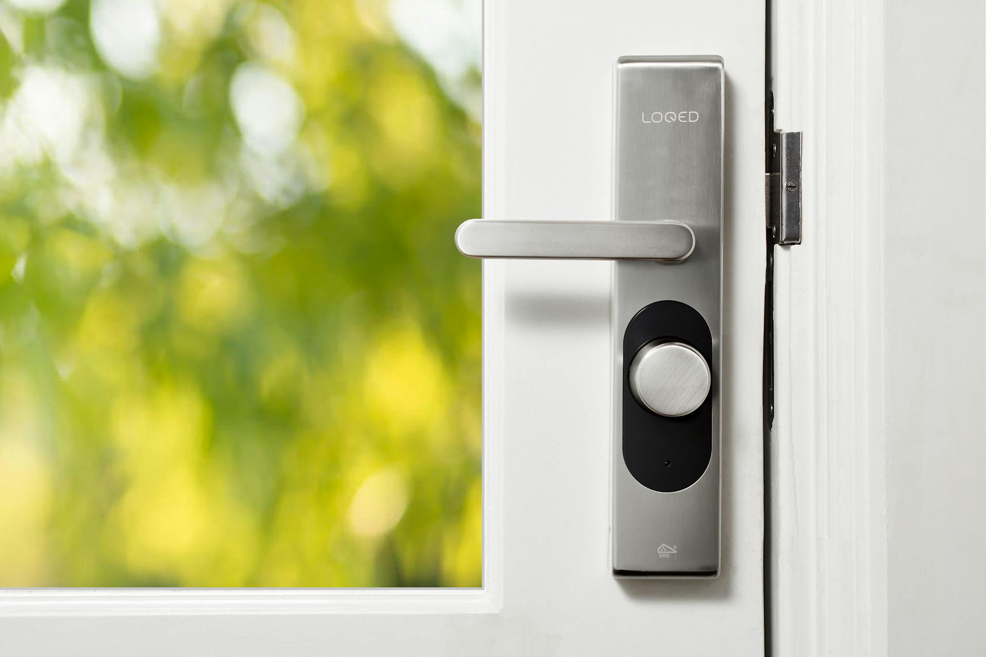 Sleek smart door lock on a white door with blurred green foliage in the background.