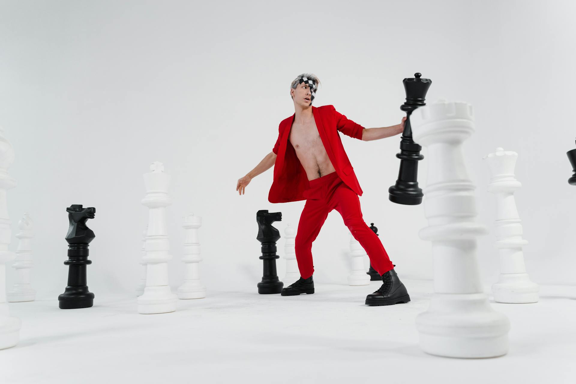Stylish man in red suit posing with oversized chess pieces in a studio setting.