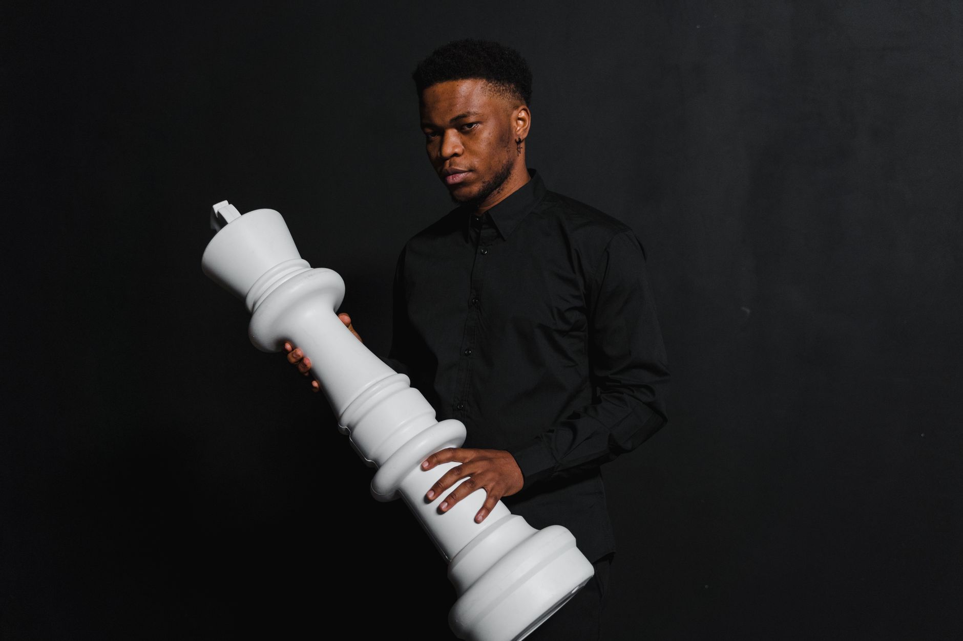 Portrait of a man in black attire holding a large white chess piece against a dark backdrop.