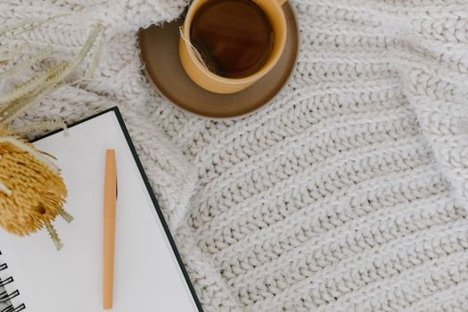A warm flat lay of a cup of tea, notebook, and pen on a knitted blanket for a cozy ambiance.