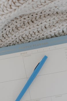 A blue pen rests on an open weekly planner against a soft, knitted backdrop, offering a cozy planning theme.