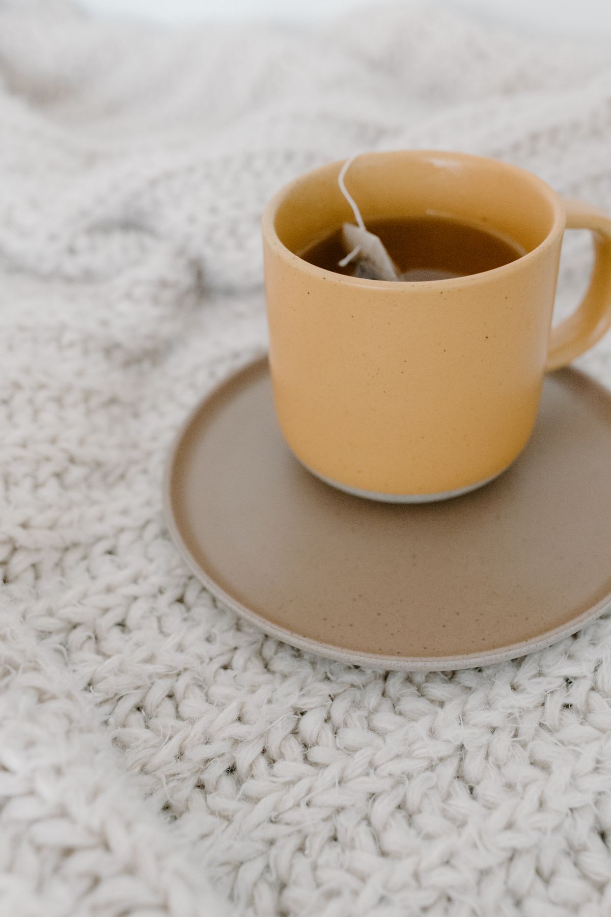A white tea cup resting on a chunky, textured white knitted blanket, the kind of wool layer that makes a sofa feel warm