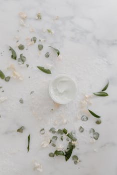 Flat lay of skincare cream on marble with green leaves, promoting natural beauty.