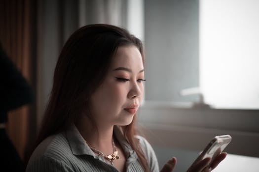 Young Asian woman using smartphone indoors, focused on screen.