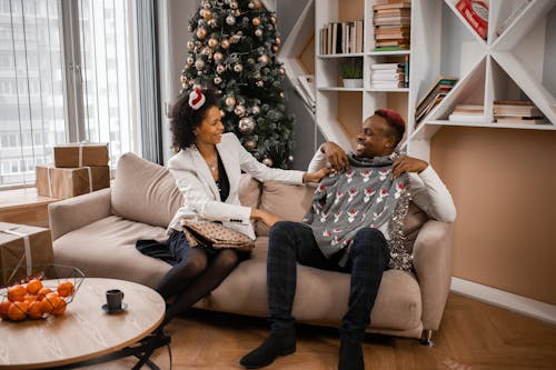 A Man Sitting on a Couch Holding an Ugly Christmas Sweater Beside a Woman