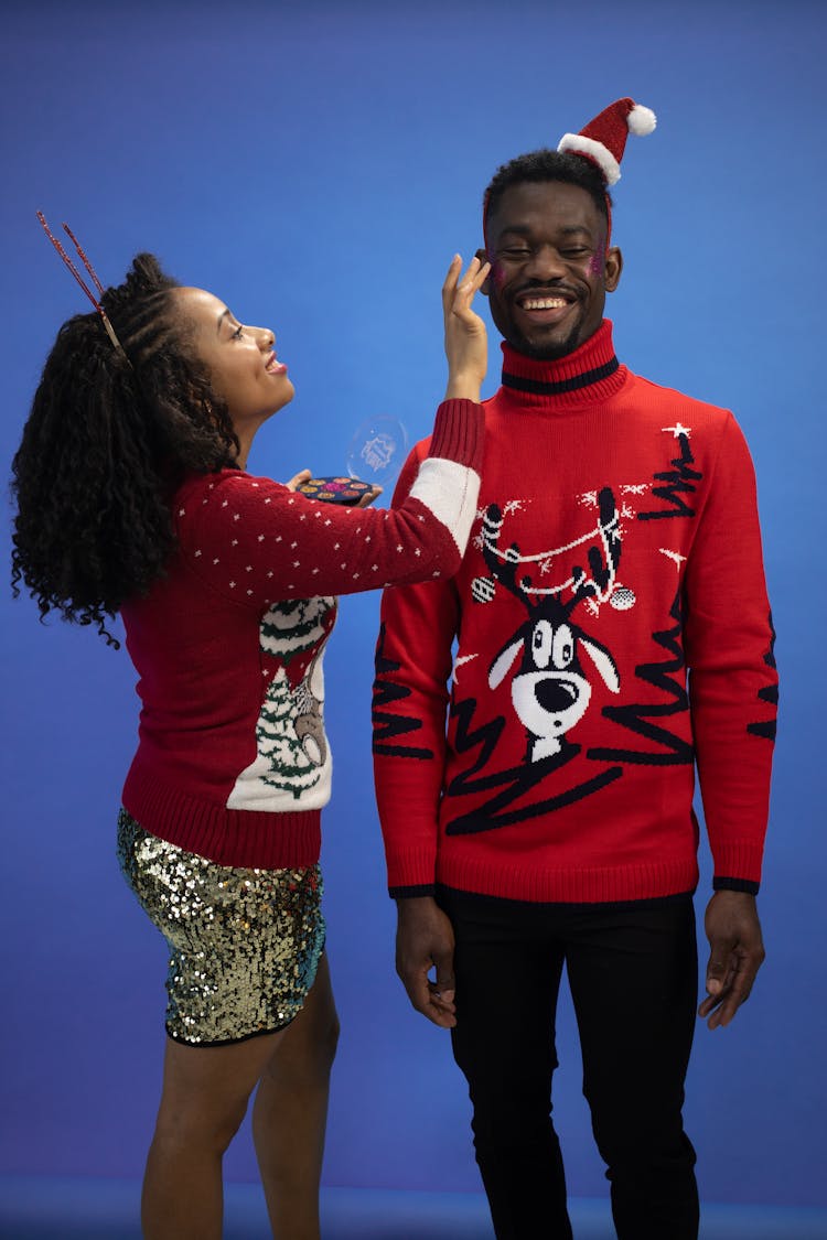 A Woman Applying Blush On On A Man Wearing An Ugly Christmas Sweater