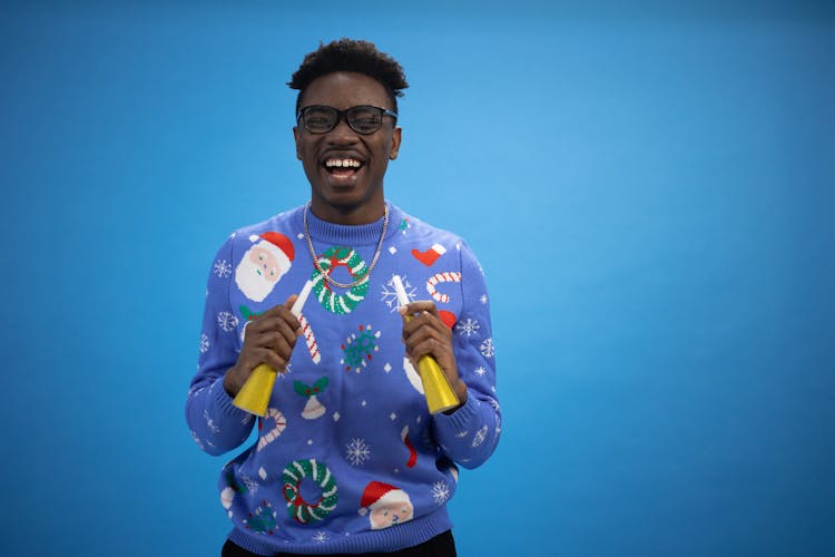 A Man Wearing A Blue Ugly Christmas Sweater Holding Toy Trumpets