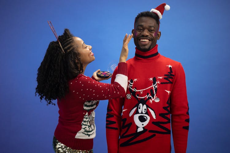 A Woman Applying Purple Blush On On A Man In Red Ugly Christmas Sweater