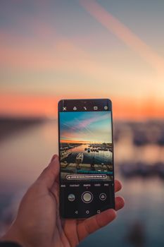 Crop anonymous person with cellphone demonstrating photo of city harbor under colorful cloudy sky at sundown