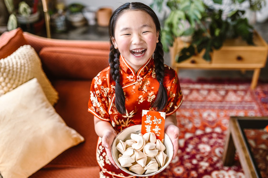 Young girl in red traditional dress holding a bowl of fortune cookies and a red envelope.