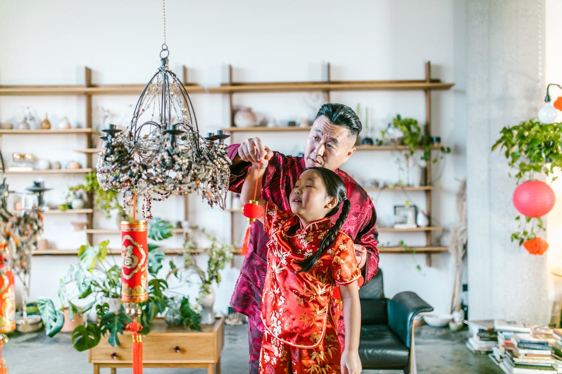 Grandfather in traditional attire helping his granddaughter hang a red lantern, sharing a joyful moment