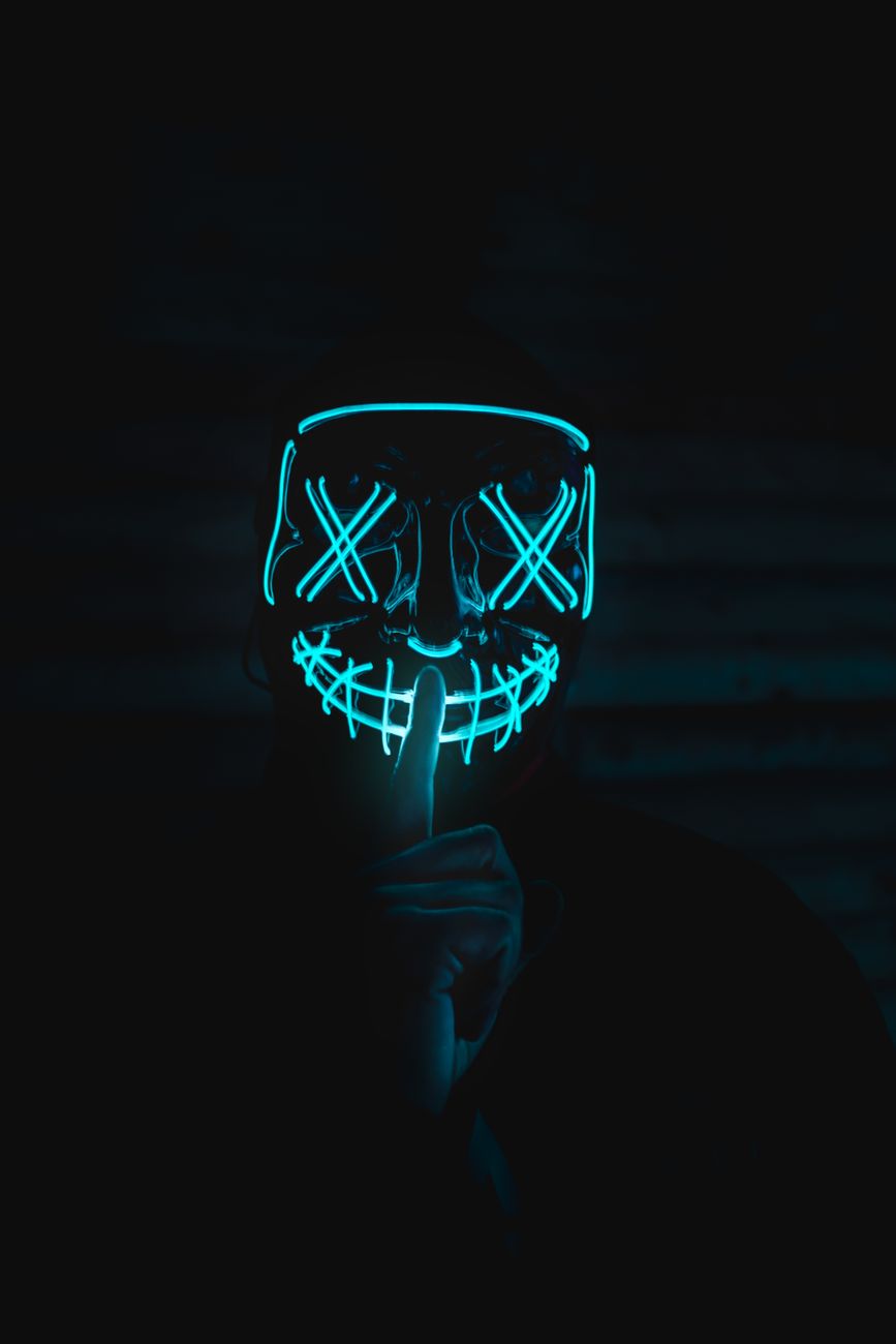 A person wearing an illuminated neon mask gesturing for silence in a dark setting.. person, wearing, illuminated