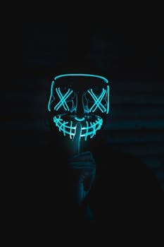 A person wearing an illuminated neon mask gesturing for silence in a dark setting.