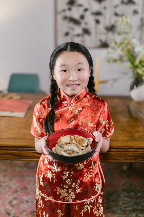 Free Young girl in red traditional dress holding a bowl of dumplings indoors. Stock Photo