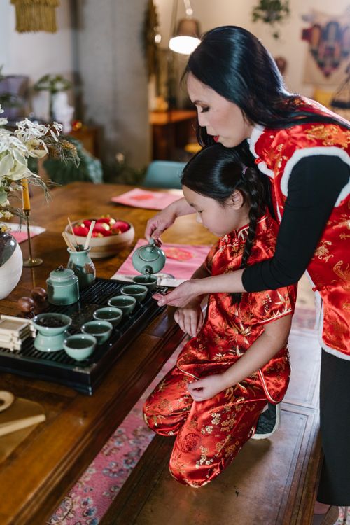 Free An Asian mother and daughter wearing traditional red Chinese clothing, pouring tea in an indoor setting. Stock Photo