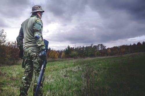 Pria Dalam Setelan Tentara Kamuflase Sambil Memegang Senapan Berburu Hitam